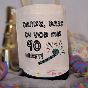 May include: A white fabric bag with a drawstring closure. The bag has black text that says "Danke. Dass du vor mir 40 wirst!" with a teal party blower graphic.
