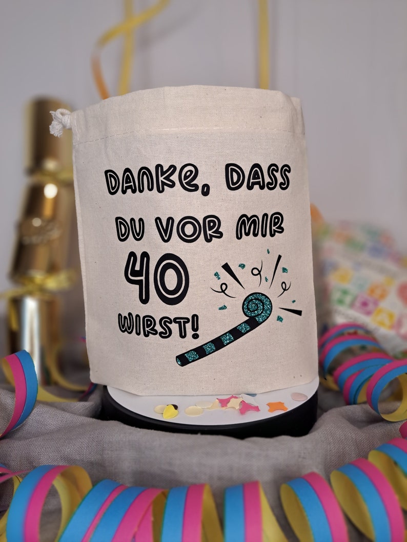 May include: A white fabric bag with a drawstring closure. The bag has black text that says "Danke. Dass du vor mir 40 wirst!" with a teal party blower graphic.