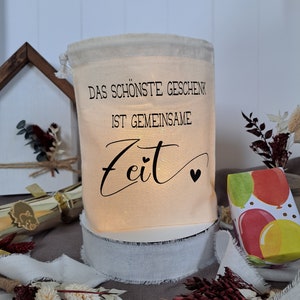 May include: A white fabric bag with a drawstring closure. The bag has black text that reads "Das sch&ouml;nste Geschenk ist gemeinsame Zeit" with two small hearts. The bag is sitting on a white base and a white ribbon.