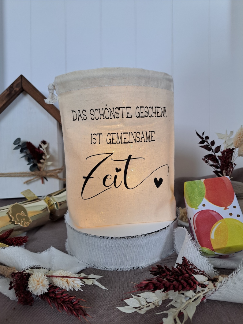 May include: A white fabric bag with a drawstring closure. The bag has black text that reads "Das sch&ouml;nste Geschenk ist gemeinsame Zeit" with two small hearts. The bag is sitting on a white base and a white ribbon.