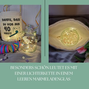 May include: A white fabric bag with the text "DANKE, DASS DU VOR MIR 40 WIRST!" printed on it. A clear glass jar with a string of fairy lights inside is sitting on a table next to the bag. The bag is open and the fairy lights are visible inside.