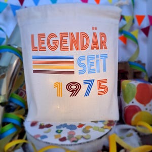 May include: A white canvas bag with the text "LEGEND&Auml;R SEIT 1975" in colorful, retro-style lettering. The bag is surrounded by colorful confetti and streamers.