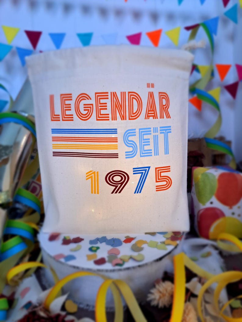 May include: A white canvas bag with the text "LEGEND&Auml;R SEIT 1975" in colorful, retro-style lettering. The bag is surrounded by colorful confetti and streamers.