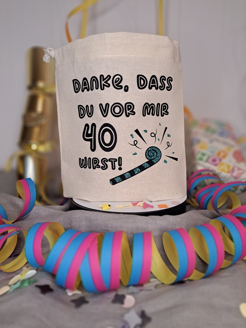 May include: A white fabric bag with a drawstring closure. The bag has a black graphic that says "Danke. Dass du vor mir 40 wirst!" with a blue and black party blower graphic.