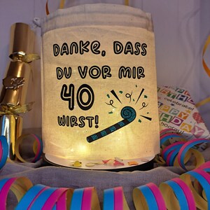 May include: A white fabric bag with a drawstring closure and a light inside. The bag has a black graphic that says "Danke, dass du vor mir 40 wirst!" with a party blower and confetti.
