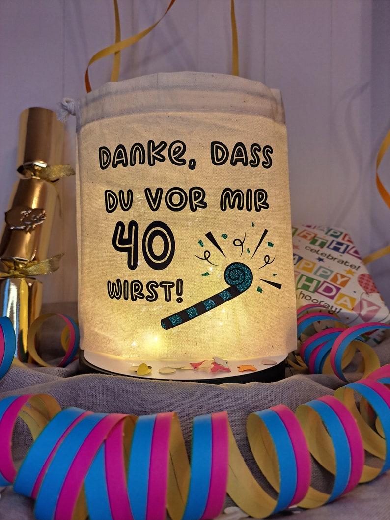 May include: A white fabric bag with a drawstring closure and a light inside. The bag has a black graphic that says "Danke, dass du vor mir 40 wirst!" with a party blower and confetti.