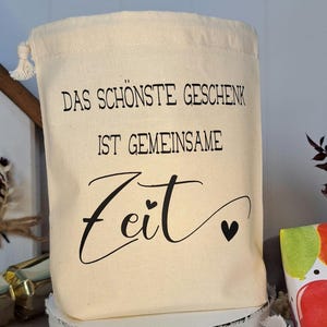 May include: A white cotton drawstring bag with the text "Das sch&ouml;nste Geschenk ist gemeinsame Zeit" in black lettering. Two small black hearts are on either side of the word "Zeit".