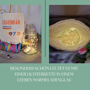 May include: A white canvas bag with the text "LEGEND&Auml;R SEIT 1973" printed on it. A clear glass jar with a string of fairy lights inside is sitting on a table next to the bag. The jar is surrounded by confetti and a colorful streamer.