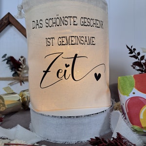 May include: A white fabric gift bag with the text "Das sch&ouml;nste Geschenk ist gemeinsame Zeit" in black lettering. The bag is lit from within and has a white ribbon around the bottom.