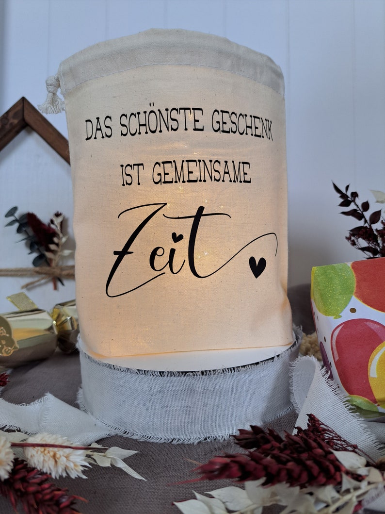 May include: A white fabric gift bag with the text "Das sch&ouml;nste Geschenk ist gemeinsame Zeit" in black lettering. The bag is lit from within and has a white ribbon around the bottom.