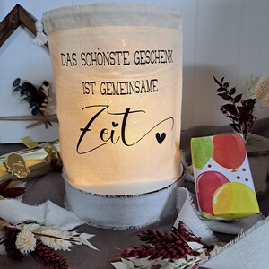 May include: A white canvas bag with a drawstring closure. The bag has a black inscription that reads "Das sch&ouml;nste Geschenk ist gemeinsame Zeit" with a small heart symbol. The bag is lit from within, creating a warm glow. The bag is sitting on a white base with a white ribbon around it. A small gift box wrapped in colorful paper with balloons is in the background.