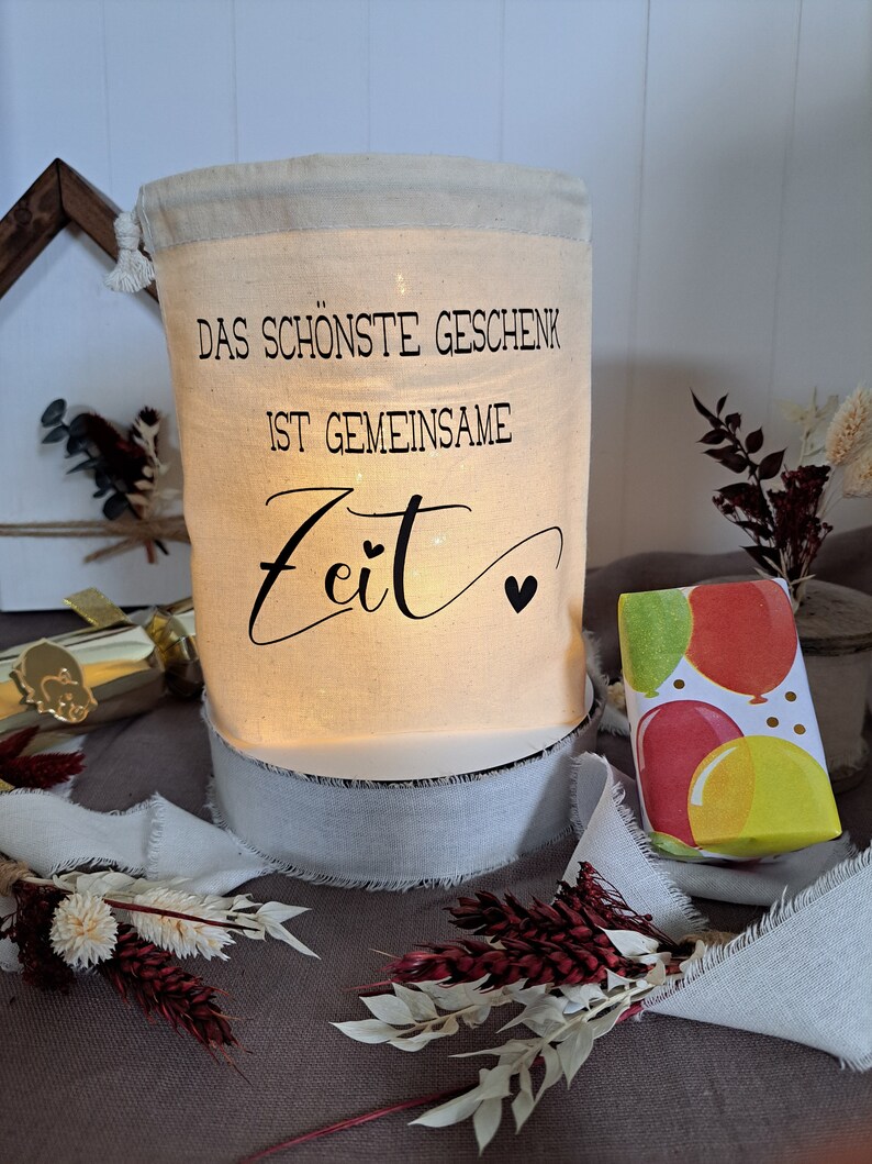 May include: A white canvas bag with a drawstring closure. The bag has a black inscription that reads "Das sch&ouml;nste Geschenk ist gemeinsame Zeit" with a small heart symbol. The bag is lit from within, creating a warm glow. The bag is sitting on a white base with a white ribbon around it. A small gift box wrapped in colorful paper with balloons is in the background.