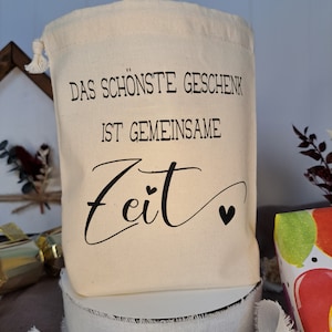 May include: A white cotton drawstring bag with black text that reads "Das sch&ouml;nste Geschenk ist gemeinsame Zeit" with a small heart symbol.