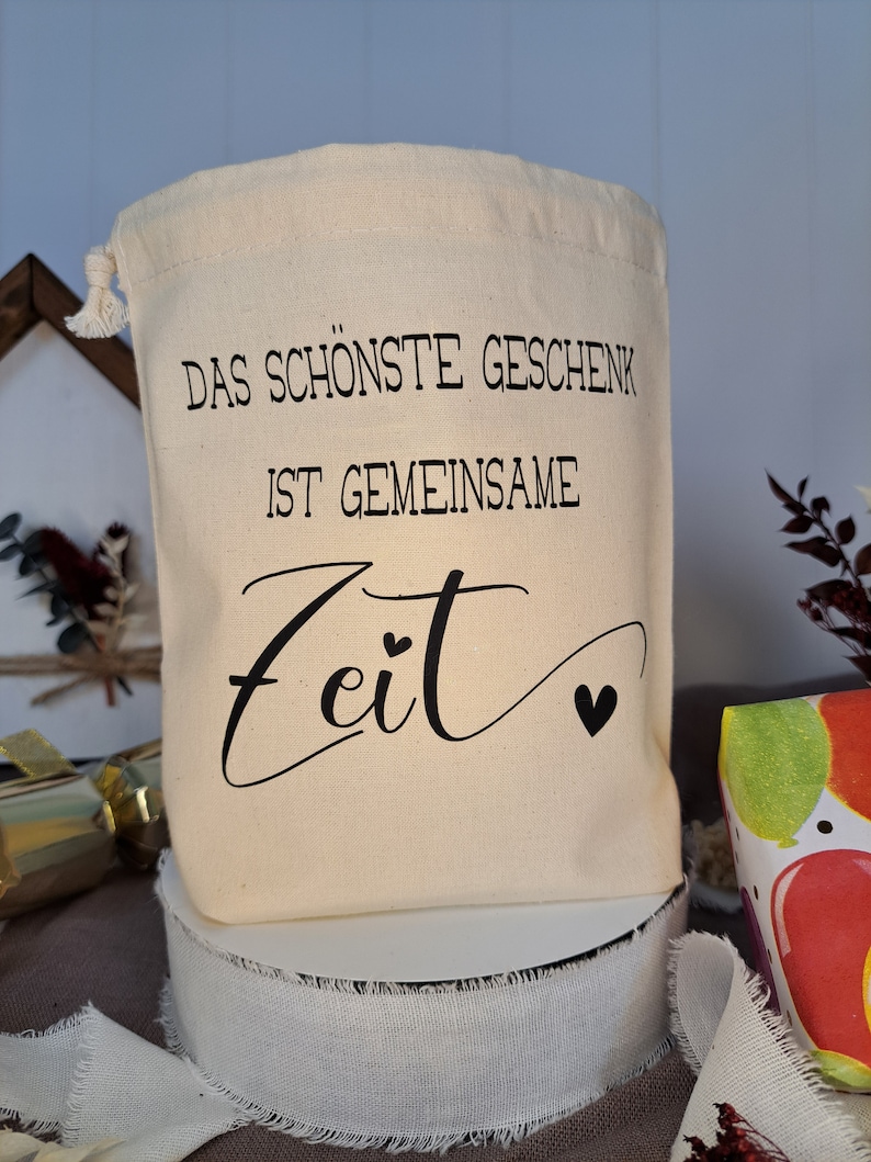 May include: A white cotton drawstring bag with black text that reads "Das sch&ouml;nste Geschenk ist gemeinsame Zeit" with a small heart symbol.