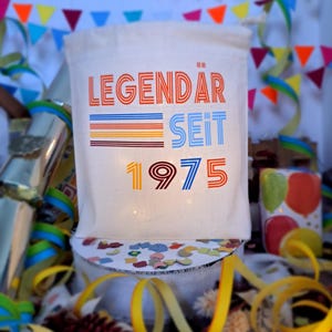 May include: A white canvas bag with the text "LEGENDAR SEIT 1975" in colorful retro-style lettering. The bag is surrounded by colorful confetti and streamers.