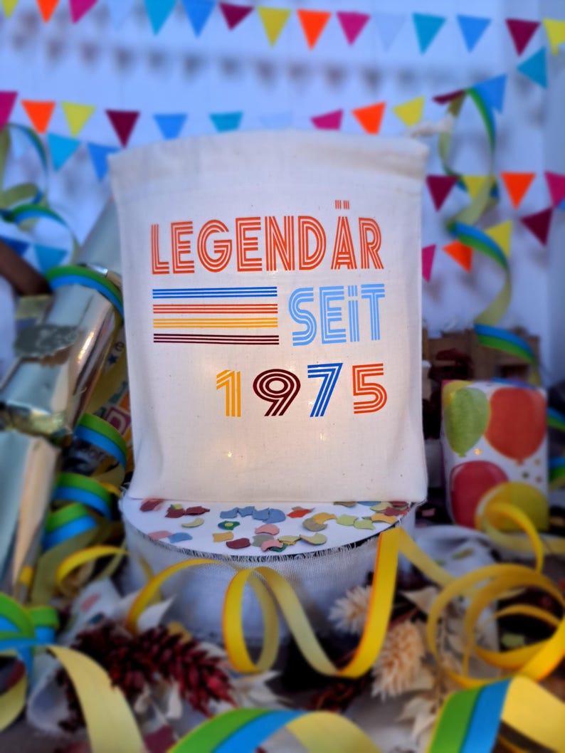 May include: A white canvas bag with the text "LEGENDAR SEIT 1975" in colorful retro-style lettering. The bag is surrounded by colorful confetti and streamers.