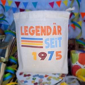 May include: A white canvas bag with the text "LEGEND&Auml;R SEIT 1975" in colorful, retro-style lettering. The bag is decorated with colorful stripes and is perfect for a birthday party.