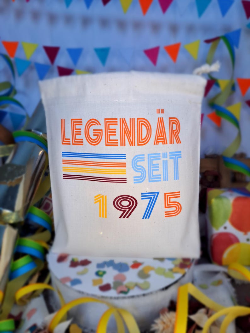 May include: A white canvas bag with the text "LEGEND&Auml;R SEIT 1975" in colorful, retro-style lettering. The bag is decorated with colorful stripes and is perfect for a birthday party.