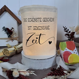 May include: A white fabric gift bag with a drawstring closure. The bag has a black print that reads "Das sch&ouml;nste Geschenk ist gemeinsame Zeit" with a small heart symbol. The bag is sitting on a white surface with a white ribbon around it.