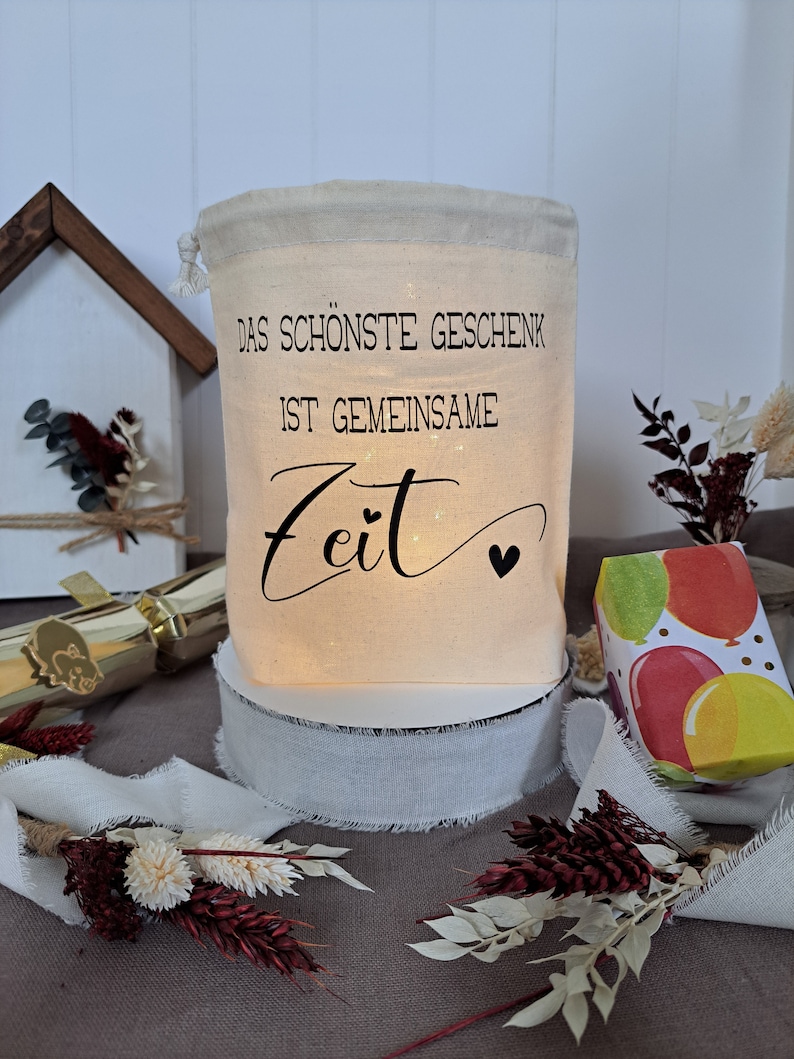 May include: A white fabric gift bag with a drawstring closure. The bag has a black print that reads "Das sch&ouml;nste Geschenk ist gemeinsame Zeit" with a small heart symbol. The bag is sitting on a white surface with a white ribbon around it.