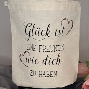 Könnte beinhalten: Eine weiße Stofftasche mit schwarzem Text, der "Glück ist eine Freundin wie dich zu haben!" lautet, mit einem Herzsymbol auf jeder Seite des Textes.