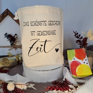 May include: A white canvas bag with a drawstring closure. The bag has black text that reads "Das sch&ouml;nste Geschenk ist gemeinsame Zeit" with a small heart symbol.