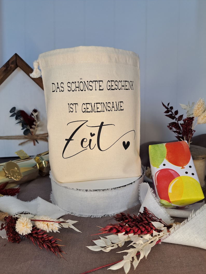 May include: A white canvas bag with a drawstring closure. The bag has black text that reads "Das sch&ouml;nste Geschenk ist gemeinsame Zeit" with a small heart symbol.