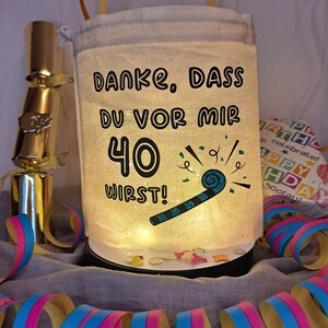 May include: A white fabric bag with a drawstring closure, decorated with a blue and black party blower and the text "DANKE, DASS DU VOR MIR 40 WIRST!" in black. The bag is lit from within, creating a warm glow.