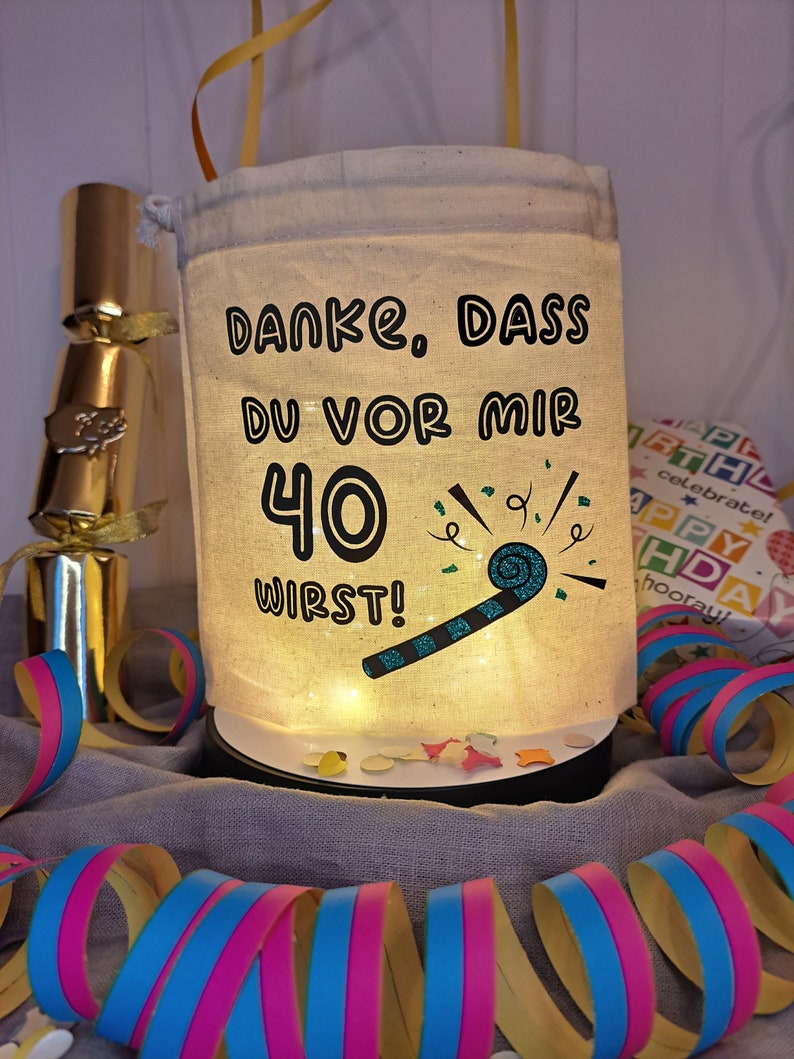 May include: A white fabric bag with a drawstring closure, decorated with a blue and black party blower and the text "DANKE, DASS DU VOR MIR 40 WIRST!" in black. The bag is lit from within, creating a warm glow.