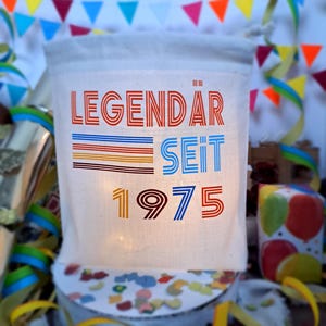 May include: A white canvas tote bag with the text "LEGENDAR SEIT 1975" in colorful retro font. The bag is decorated with colorful stripes and is illuminated from behind.