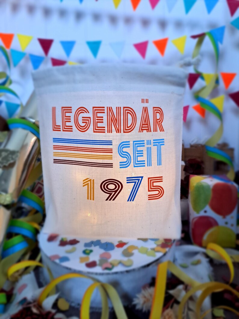 May include: A white canvas tote bag with the text "LEGENDAR SEIT 1975" in colorful retro font. The bag is decorated with colorful stripes and is illuminated from behind.