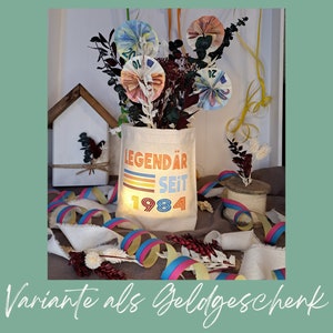 May include: A white canvas tote bag with the text "LEGEND&Auml;R SEIT 1984" printed on it in colorful retro-style lettering. The bag is filled with rolled-up bills of money, representing a creative and unique gift idea.