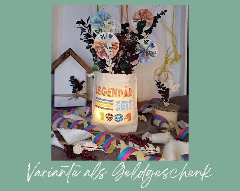 May include: A white canvas tote bag with the text "LEGEND&Auml;R SEIT 1984" printed on it in colorful retro-style lettering. The bag is filled with rolled-up bills of money, representing a creative and unique gift idea.