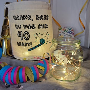 May include: A white fabric bag with the text "Danke, dass du vor mir 40 wirst!" and a green party blower graphic. The bag is sitting on a gray surface with colorful confetti and a glass jar with white fairy lights.