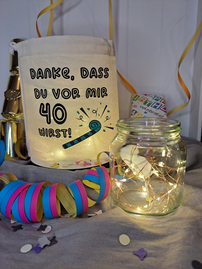 May include: A white fabric bag with the text "Danke, dass du vor mir 40 wirst!" and a green party blower graphic. The bag is sitting on a gray surface with colorful confetti and a glass jar with white fairy lights.