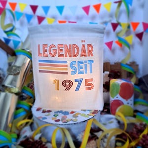 May include: A white canvas bag with the text "LEGENDAR SEIT 1975" printed in red, blue, and orange. The bag is surrounded by colorful confetti and streamers.