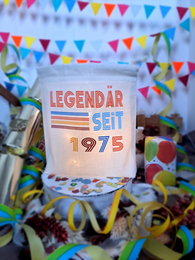 May include: A white canvas bag with the text "LEGENDAR SEIT 1975" printed in red, blue, and orange. The bag is surrounded by colorful confetti and streamers.