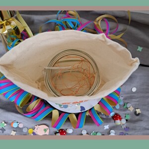 May include: A white fabric bag with a clear glass jar containing a copper wire light string inside. The bag is surrounded by colorful streamers and confetti.