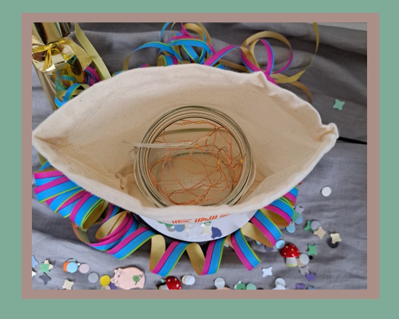May include: A white fabric bag with a clear glass jar containing a copper wire light string inside. The bag is surrounded by colorful streamers and confetti.