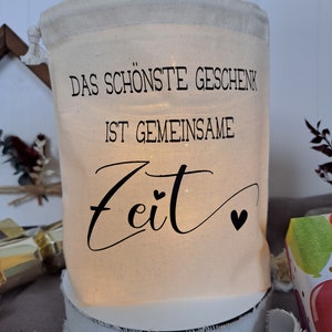 May include: A white fabric bag with a drawstring closure. The bag has a black print that reads "Das sch&ouml;nste Geschenk ist gemeinsame Zeit" with two small hearts.