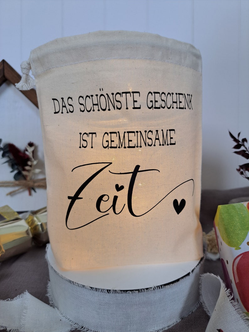 May include: A white fabric bag with a drawstring closure. The bag has a black print that reads "Das sch&ouml;nste Geschenk ist gemeinsame Zeit" with two small hearts.
