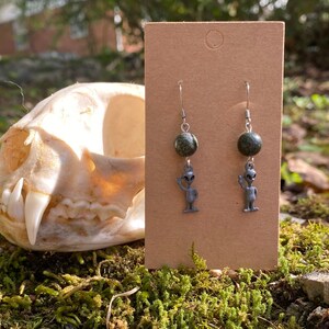 May include: A pair of silver earrings with green stone beads and silver alien charms. The earrings are displayed on a brown card.