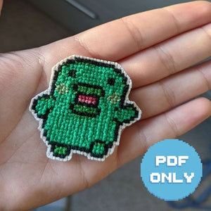 Peut inclure: Motif de point de croix vert représentant un monstre de dessin animé avec un large sourire et de grands yeux. Le motif est sur un tissu blanc et a un contour noir. Le texte "PDF ONLY" est visible dans le coin inférieur droit de l'image.