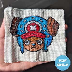 May include: A cross-stitch embroidery on light beige fabric featuring the character Tony Tony Chopper from One Piece. The design displays a tan face, a red hat with a white cross, and a blue hat cover with brown antlers, all outlined in black thread.