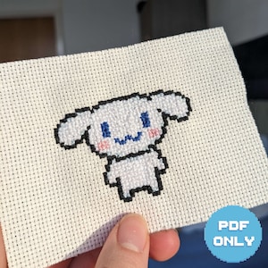 May include: White cross-stitch embroidery of a cartoon dog with blue eyes and a pink nose. The dog is smiling and has a black outline.
