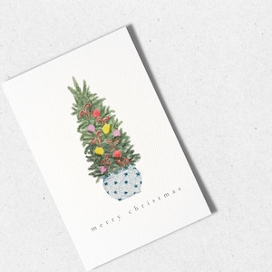 May include: A watercolor illustration of a small Christmas tree decorated with red, yellow, and pink ornaments and red bows in a blue and white patterned pot. The text "merry christmas" is written below the tree.