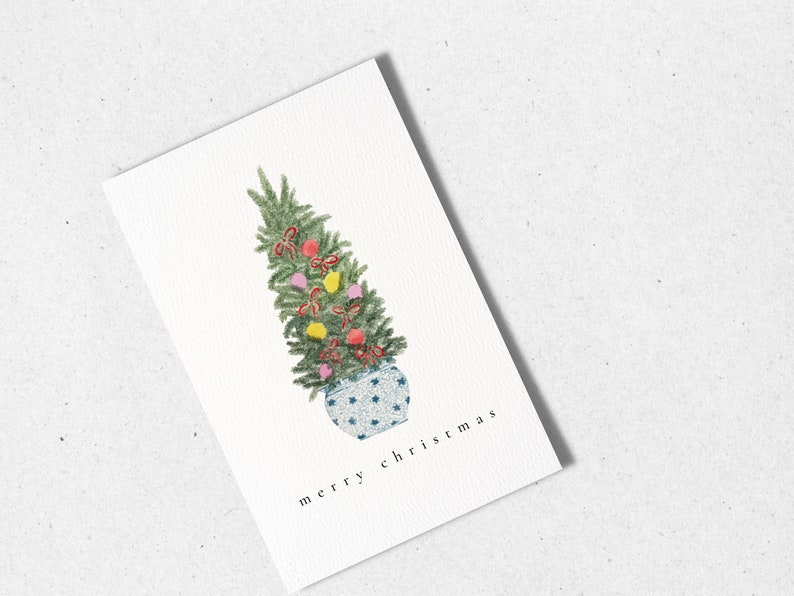 May include: A watercolor illustration of a small Christmas tree decorated with red, yellow, and pink ornaments and red bows in a blue and white patterned pot. The text "merry christmas" is written below the tree.