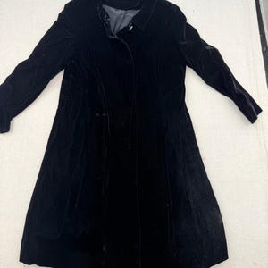 May include: A long-sleeved black velvet coat featuring a classic collar, a button-front closure, and a knee-length hemline. The garment is constructed with a soft, plush texture and includes a smooth interior lining visible at the neckline.
