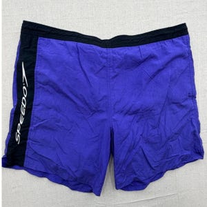 May include: Purple swim trunks with a black elastic waistband and a vertical black side panel on the left leg. The word SPEEDO is printed in white capital letters vertically along the black side panel of these athletic shorts.
