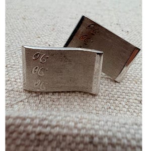 May include: A pair of rectangular silver-toned metal cufflinks with a curved surface and a fine horizontal brushed texture. Each cufflink features the number 96 embossed in a repeating vertical pattern on the left side of the face.